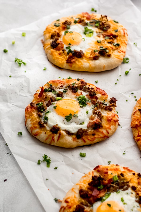 Build Your Own Breakfast Pizzas - Made with pre-made dough for ease and topped with your favorite toppings. We love sausage, bacon, a soft egg, green onion, and extra cheese. It's a brunch party, and you're all invited! #breakfastrecipes #breakfastpizza #pizzarecipes #homemadepizza #brunchrecipes #baconrecipes #bluebowlrecipes | bluebowlrecipes.com Breakfast pizzas on a white surface.