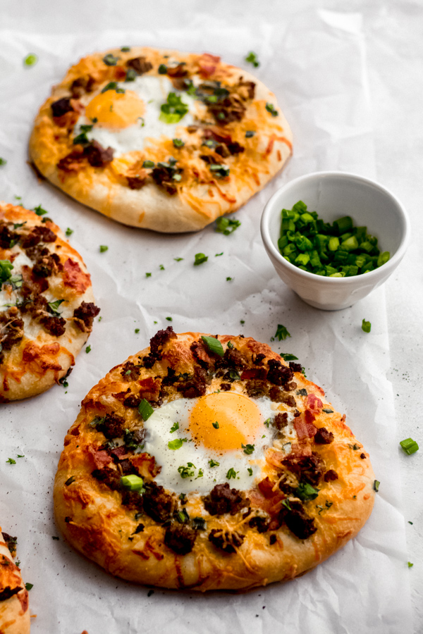 Build Your Own Breakfast Pizzas - Made with pre-made dough for ease and topped with your favorite toppings. We love sausage, bacon, a soft egg, green onion, and extra cheese. It's a brunch party, and you're all invited! #breakfastrecipes #breakfastpizza #pizzarecipes #homemadepizza #brunchrecipes #baconrecipes #bluebowlrecipes | bluebowlrecipes.com Breakfast pizzas on a white surface.