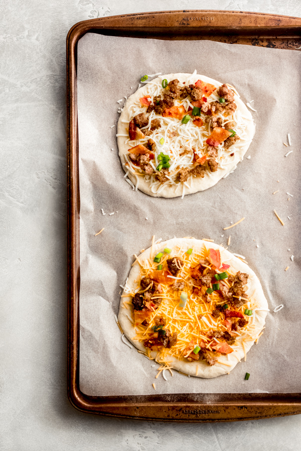 Build Your Own Breakfast Pizzas - Made with pre-made dough for ease and topped with your favorite toppings. We love sausage, bacon, a soft egg, green onion, and extra cheese. It's a brunch party, and you're all invited! #breakfastrecipes #breakfastpizza #pizzarecipes #homemadepizza #brunchrecipes #baconrecipes #bluebowlrecipes | bluebowlrecipes.com Breakfast pizzas assembled on a baking tray.