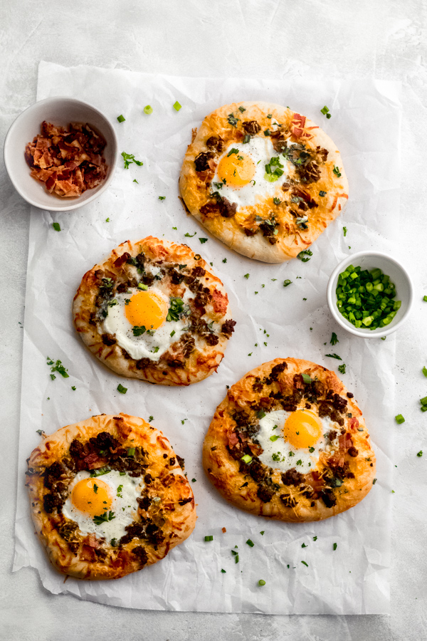 Build Your Own Breakfast Pizzas - Made with pre-made dough for ease and topped with your favorite toppings. We love sausage, bacon, a soft egg, green onion, and extra cheese. It's a brunch party, and you're all invited! #breakfastrecipes #breakfastpizza #pizzarecipes #homemadepizza #brunchrecipes #baconrecipes #bluebowlrecipes | bluebowlrecipes.com Breakfast pizzas on a white surface, garnished with parsley.