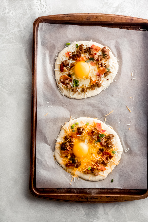 Build Your Own Breakfast Pizzas - Made with pre-made dough for ease and topped with your favorite toppings. We love sausage, bacon, a soft egg, green onion, and extra cheese. It's a brunch party, and you're all invited! #breakfastrecipes #breakfastpizza #pizzarecipes #homemadepizza #brunchrecipes #baconrecipes #bluebowlrecipes | bluebowlrecipes.com Breakfast pizzas assembled on a baking tray with eggs added.