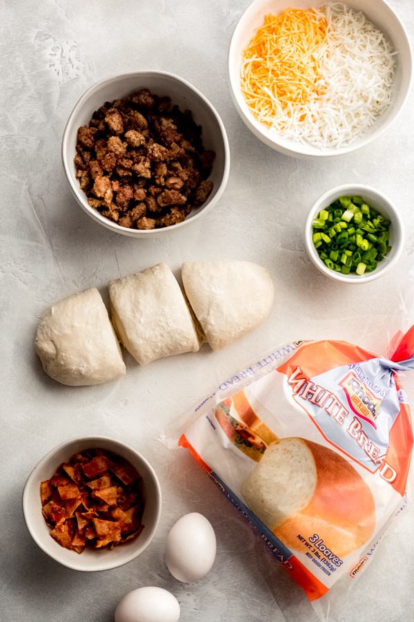 Build Your Own Breakfast Pizzas - Made with pre-made dough for ease and topped with your favorite toppings. We love sausage, bacon, a soft egg, green onion, and extra cheese. It's a brunch party, and you're all invited! #breakfastrecipes #breakfastpizza #pizzarecipes #homemadepizza #brunchrecipes #baconrecipes #bluebowlrecipes | bluebowlrecipes.com Breakfast pizza ingredients in bowls.