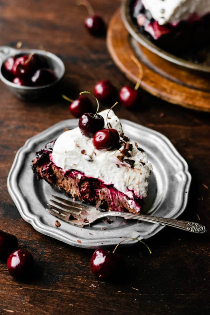 A slice of Black Forest Pie on a plate, with a fork and fresh cherries scattered around.