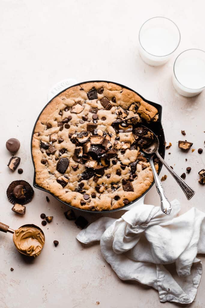 The skillet cookie topped with a pile of chopped peanut butter cups and chocolate chips, with spoons digging in.