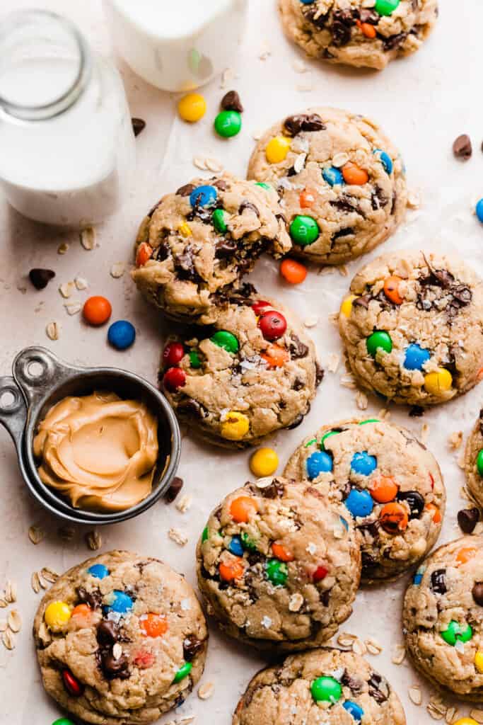 Jumbo monster cookies on a light surface, with glasses of milk and a measuring cup of peanut butter.
