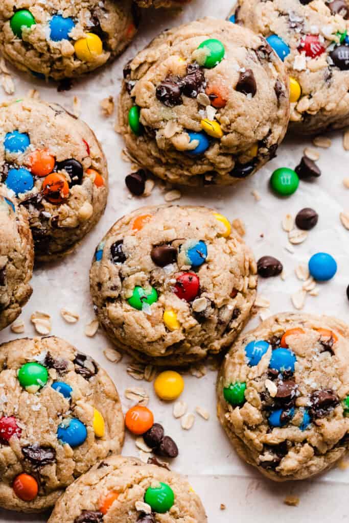 Monster cookies on a light surface with m&ms, chocolate chips, and oats sprinkled around.