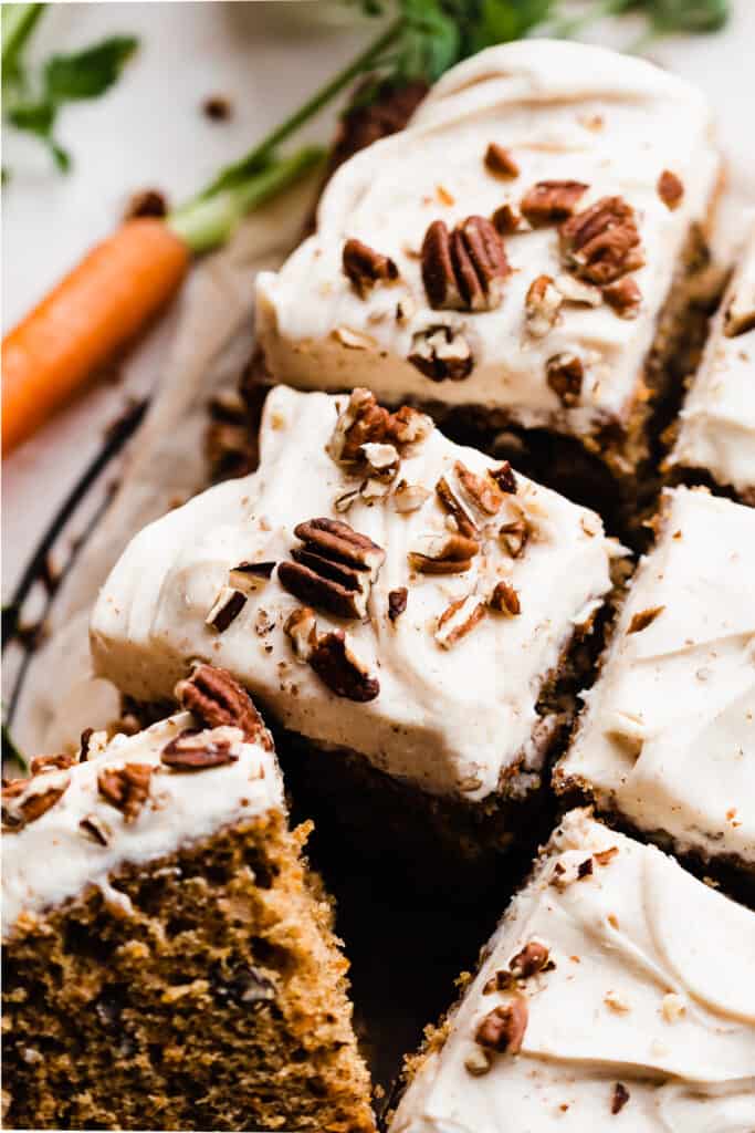 A close-up of squares of carrot cake with cream cheese frosting and chopped nuts on top.