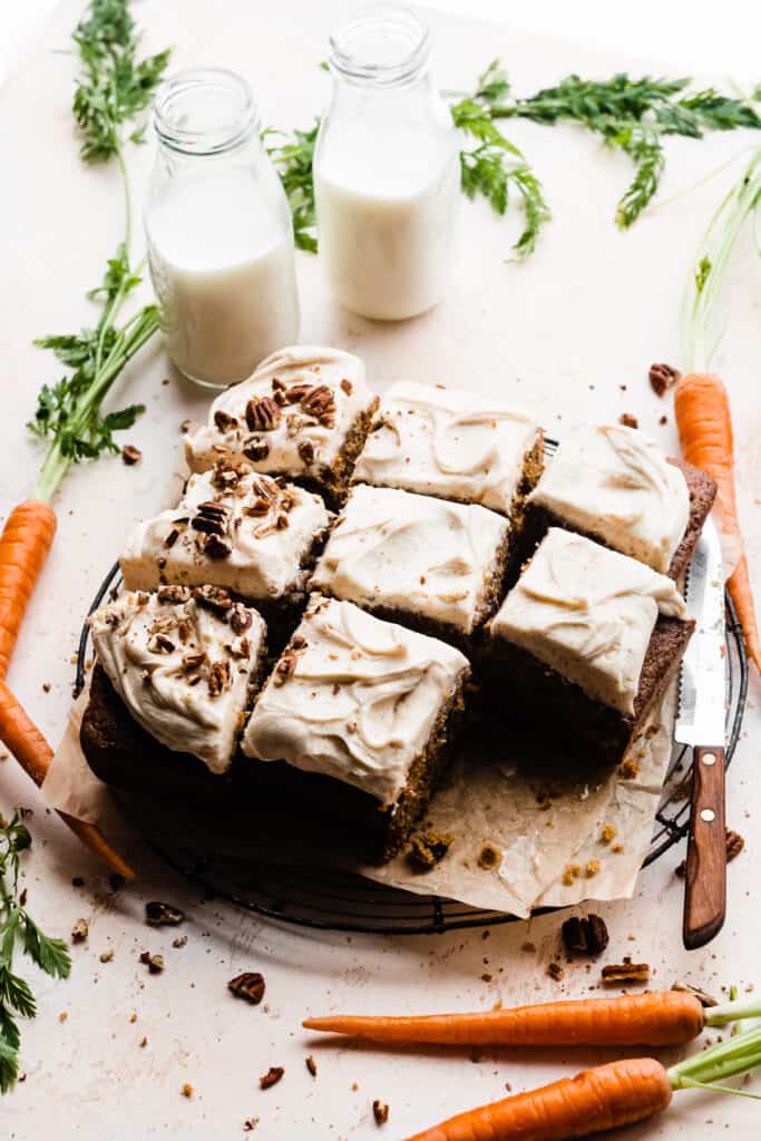 The full sliced carrot cake, topped with crushed pecans and surrounded by carrots and bottles of milk.