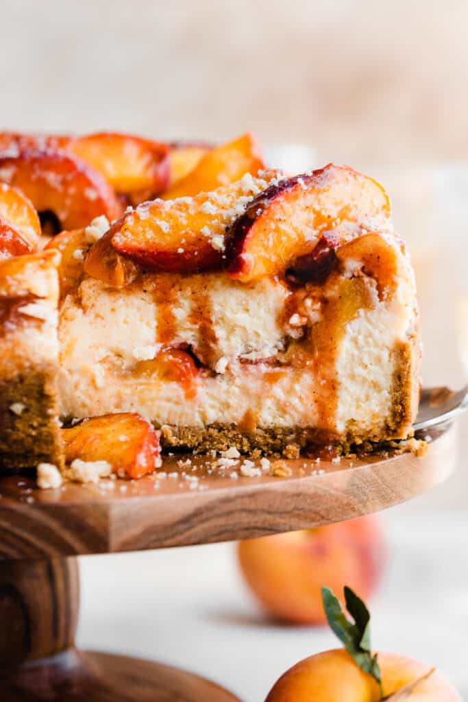 A close-up of a cheesecake slice with peaches in the center and on top, and cinnamon peach juice dripping down.