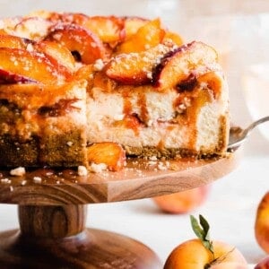 The peach cobbler cheesecake on a cake stand with a cross section cut. Juicy peach slices are on top with cinnamon peach juice dripping down.