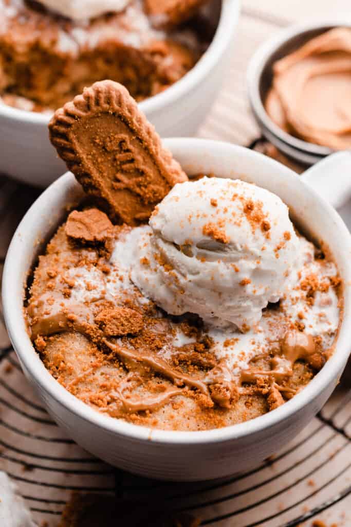 A close-up of a mug of cookie butter cake topped with cookie butter drizzle, cookie crumbs, and vanilla ice cream.