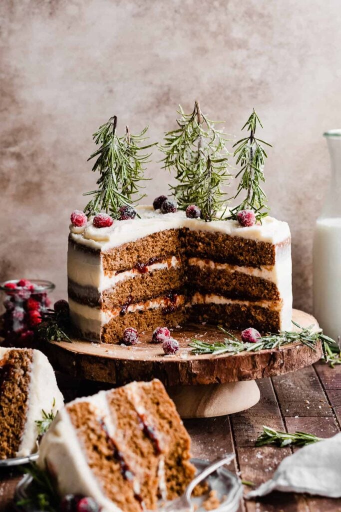 A sliced open gingerbread cake on a wooden stand, with rosemary trees and sugared cranberries.