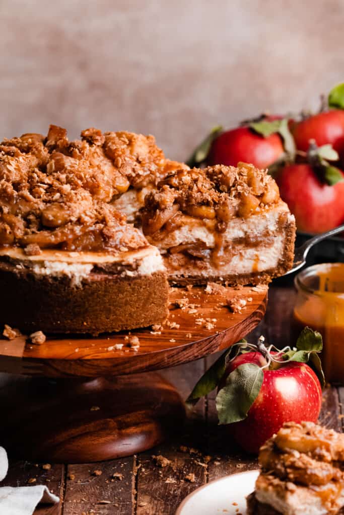A slice of the apple pie cheesecake being lifted out of the sliced cheesecake on a cake stand.