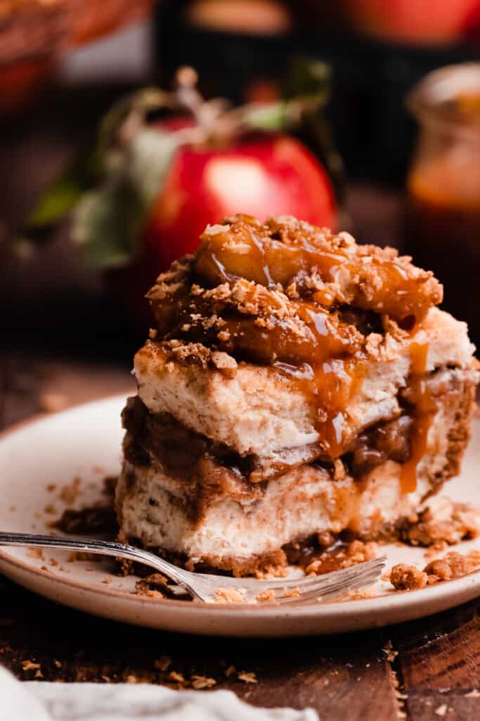 A close-up on a slice of apple cheesecake on a plate, with a bite missing. It's topped with thick caramelized apples, streusel, & caramel drizzle.