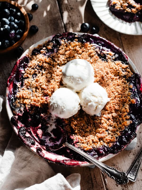 A round dish of blueberry crisp topped with ice cream, with a serving spoon digging in.