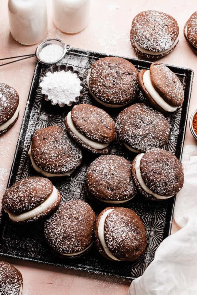 Assembled chocolate whoopie pies dusted with powdered sugar, on a vintage cookie sheet on a light pink background.