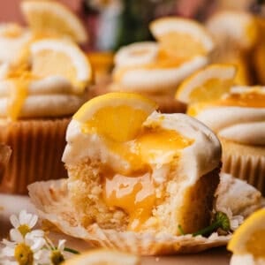 A lemon cupcake on a marble cake plate, surrounded by more lemon cupcakes. The one in focus has bites missing, to show the lemon curd filling inside.
