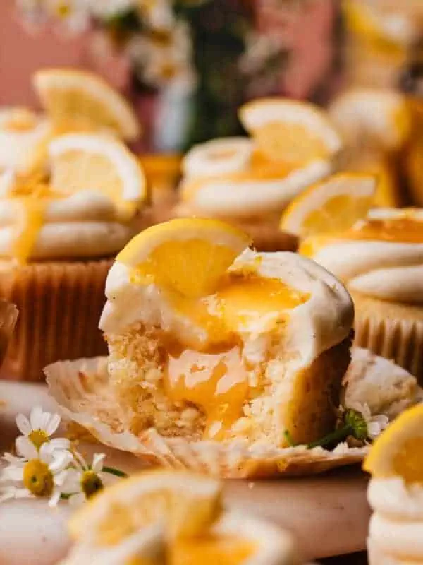 A lemon cupcake on a marble cake plate, surrounded by more lemon cupcakes. The one in focus has bites missing, to show the lemon curd filling inside.
