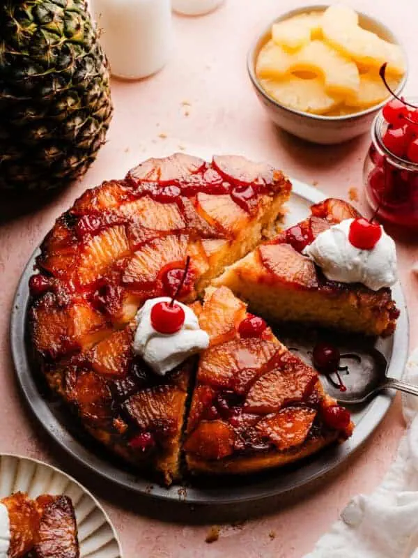 The sliced pineapple upside down cake on a cake plate. Slices are topped with dollops of whipped cream and maraschino cherries.