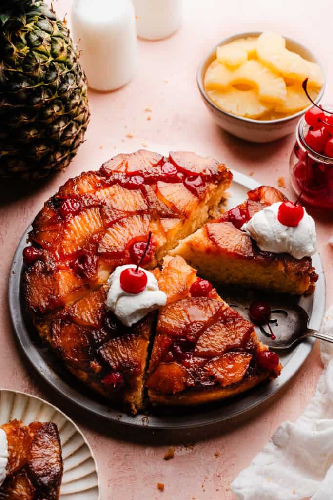 The pineapple upside down cake sliced, on a cake plate. A few of the slices have dollops of whipped cream and a maraschino cherry on top.