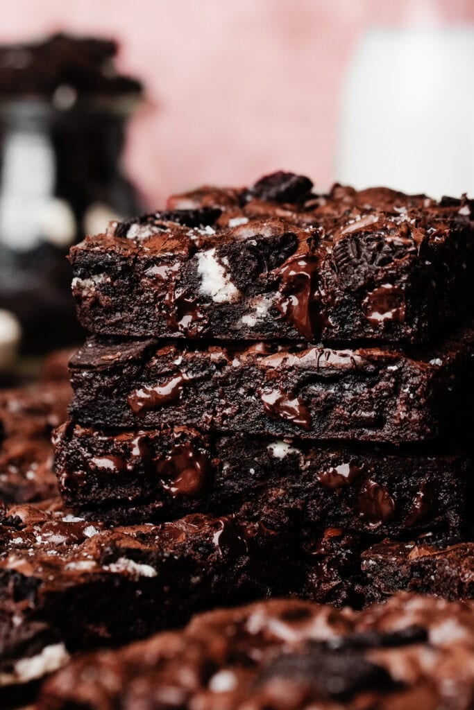 A stack of three fudgy oreo brownies, with a jar of oreo pieces and a bottle of milk in the background.