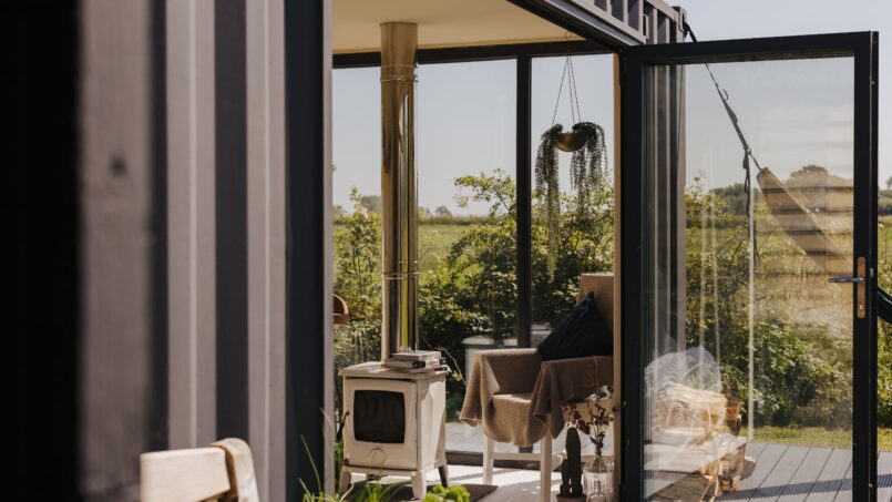 Cozy off-grid tiny home interior with large glass doors, lush outdoor view, wood-burning stove, and eco-friendly decor, perfect for sustainable living and off-grid lifestyles.