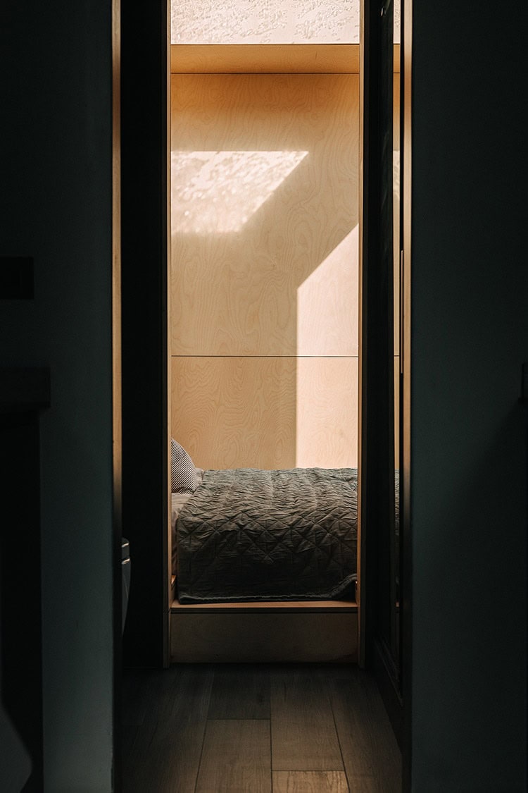 Small cozy bedroom with natural wood accent wall and sunlight streaming through window, minimalist off-grid living space, sustainable home design, eco-friendly bedroom decor.