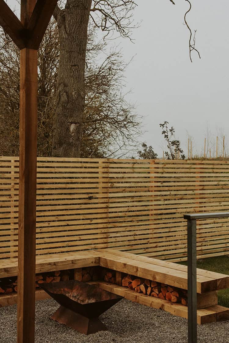Fire pit with wood storage on a deck in a peaceful garden setting.