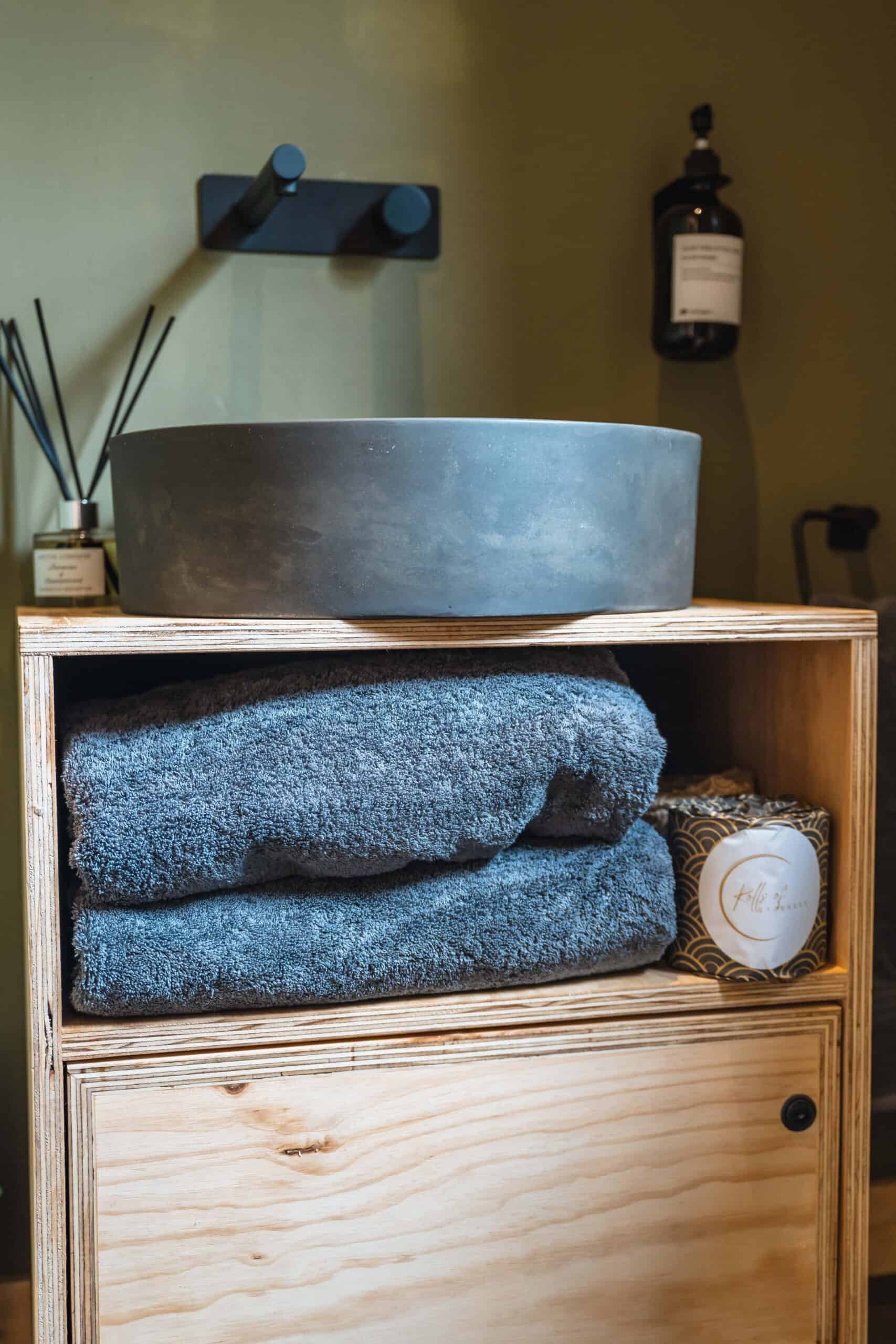 Soft grey towels and a black stone basin on a wooden shelf, accented by minimalist decor in a cozy off-grid cabin bathroom.