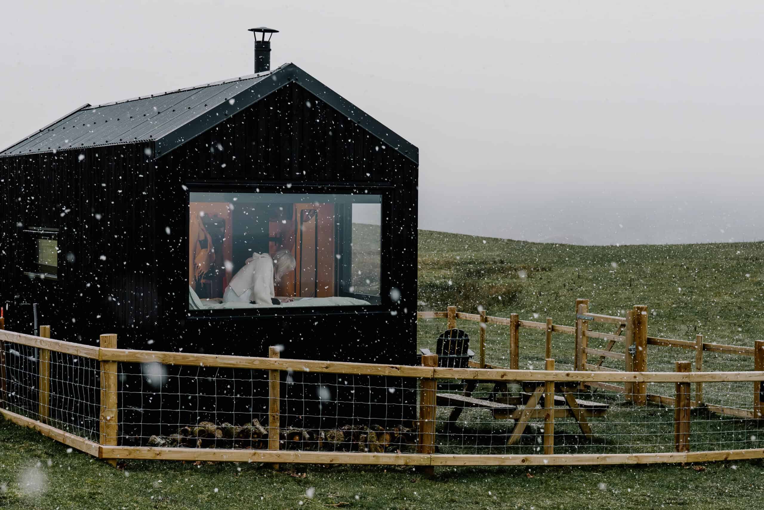 Cozy off grid tiny house during snowfall on a rural hillside, with a wooden fence and outdoor seating, perfect for a sustainable escape in nature.