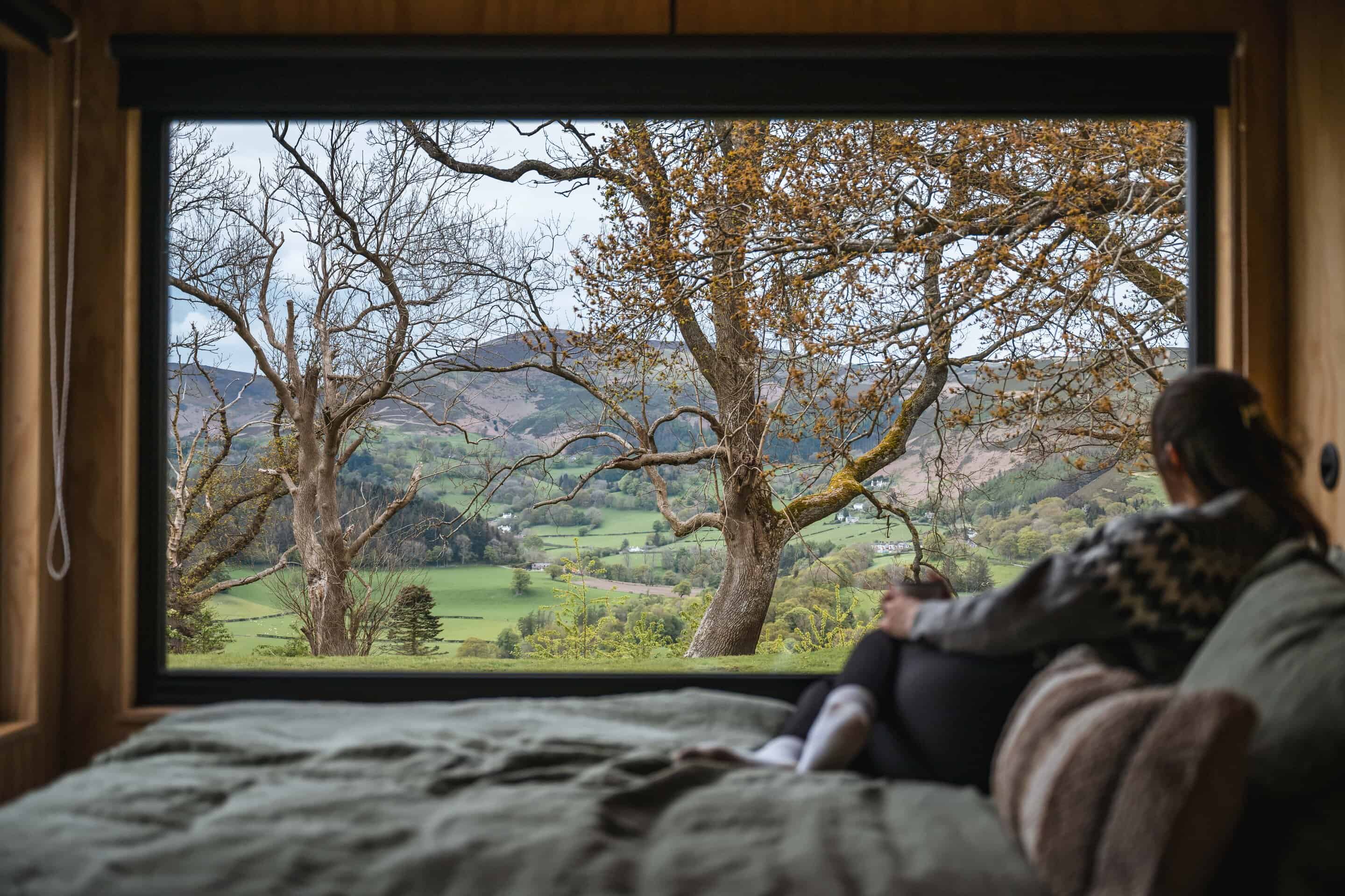 Secluded off-grid cabin view with large window overlooking rural landscape and trees in spring, eco-friendly sustainable living, peaceful nature retreat for off-grid getaway, cosy interior with forest scenery.