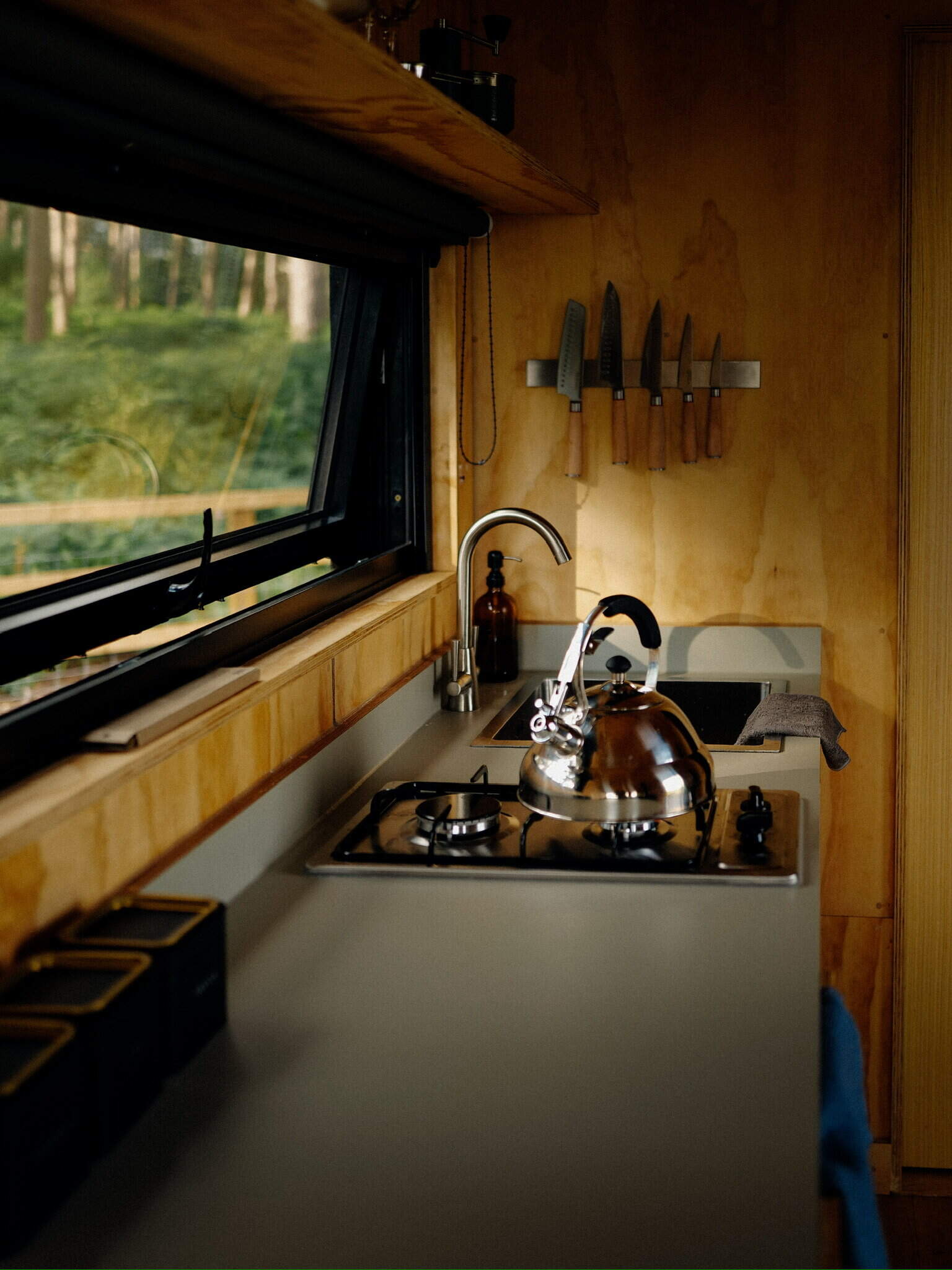 Eco-friendly off grid kitchen with wood paneling, gas stove, stainless steel kettle, and knives on wall, offering sustainable living experience in a tiny home surrounded by nature.