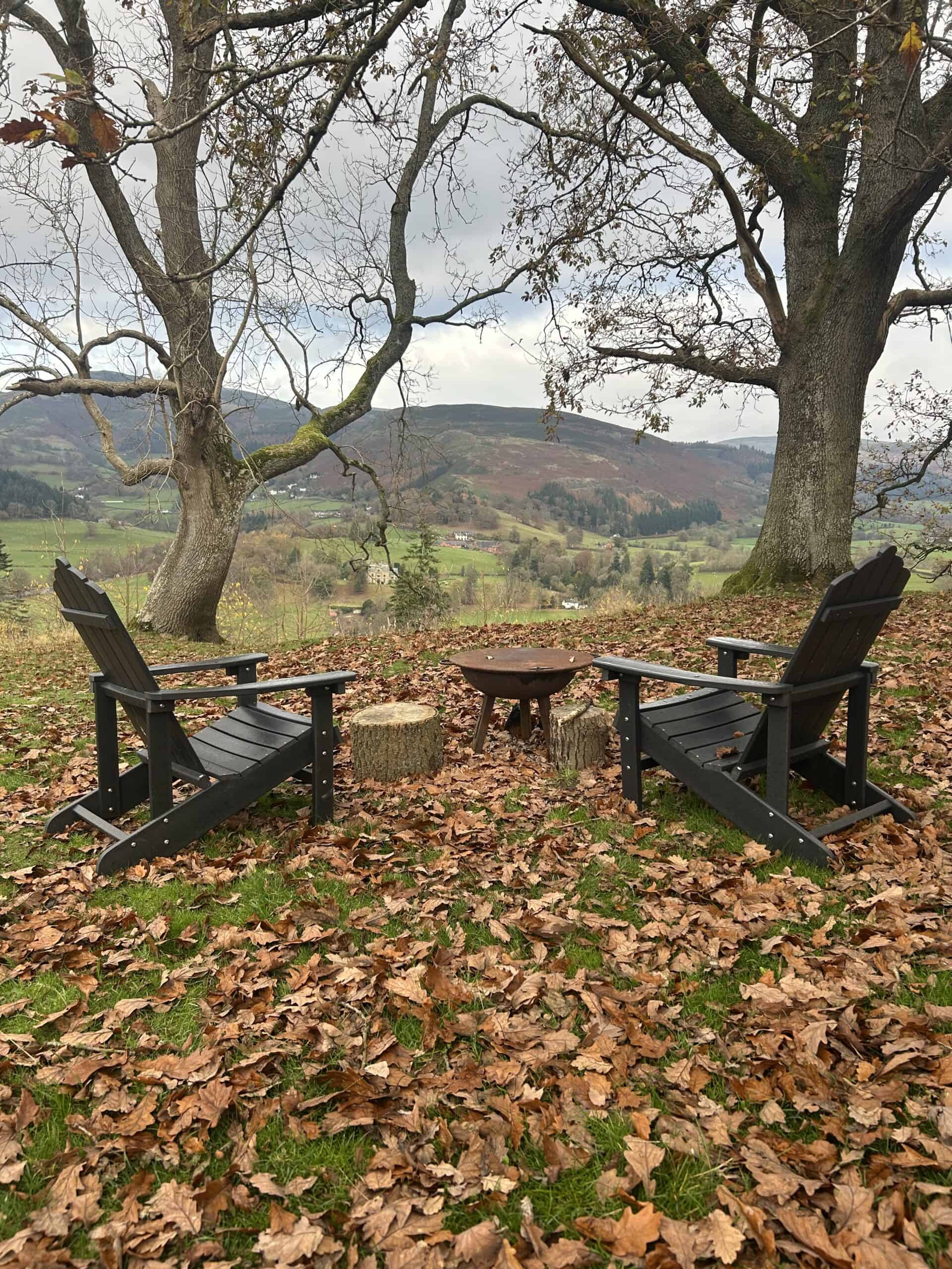 Secluded off grid camping spot with black wooden chairs and fire pit under large trees in autumn, mountain views, peaceful outdoor wilderness retreat, perfect for off grid living, nature escape.