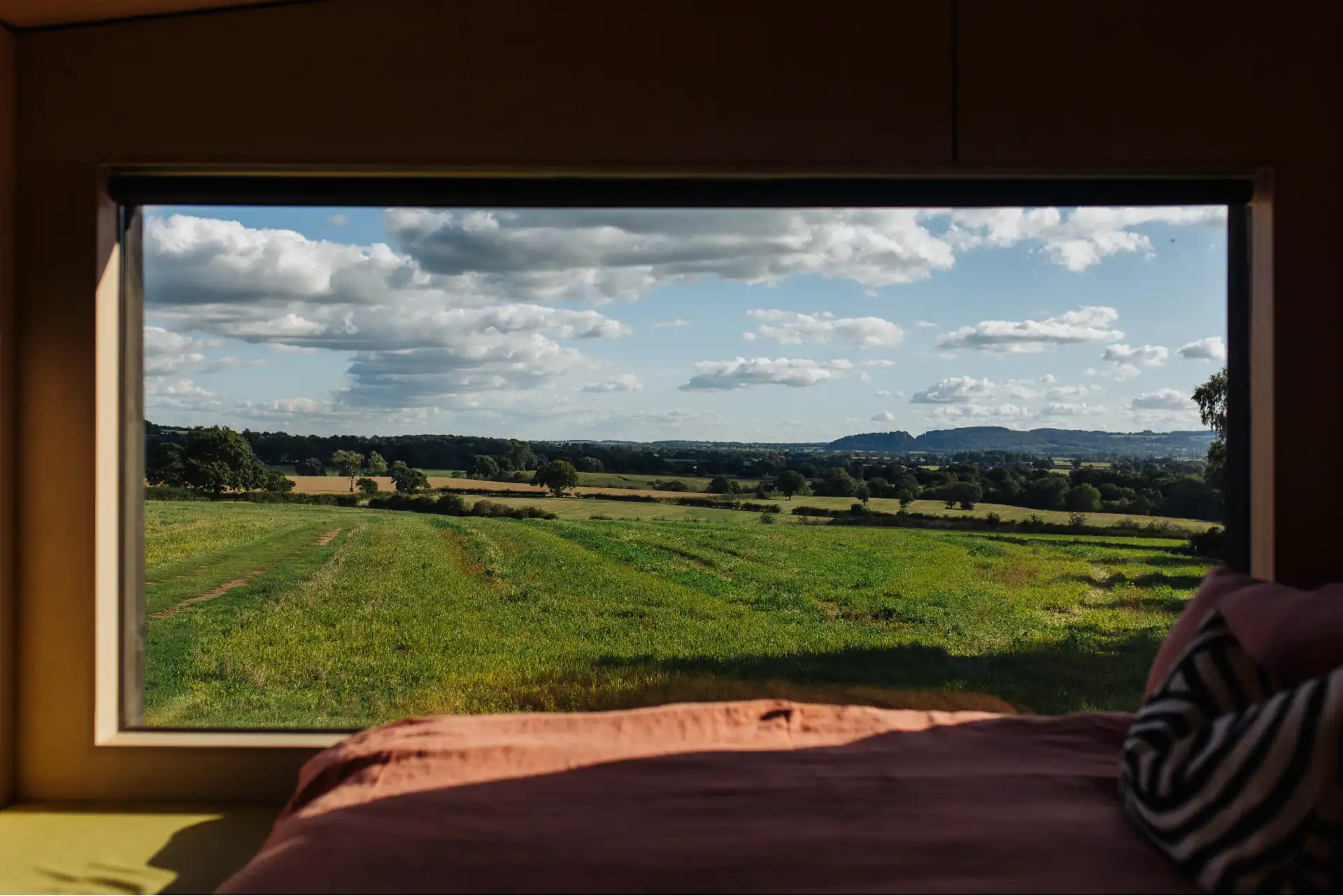 Serene countryside view through a large window, showcasing open fields, rolling hills, and a partly cloudy sky, perfect for a off-grid living experience.
