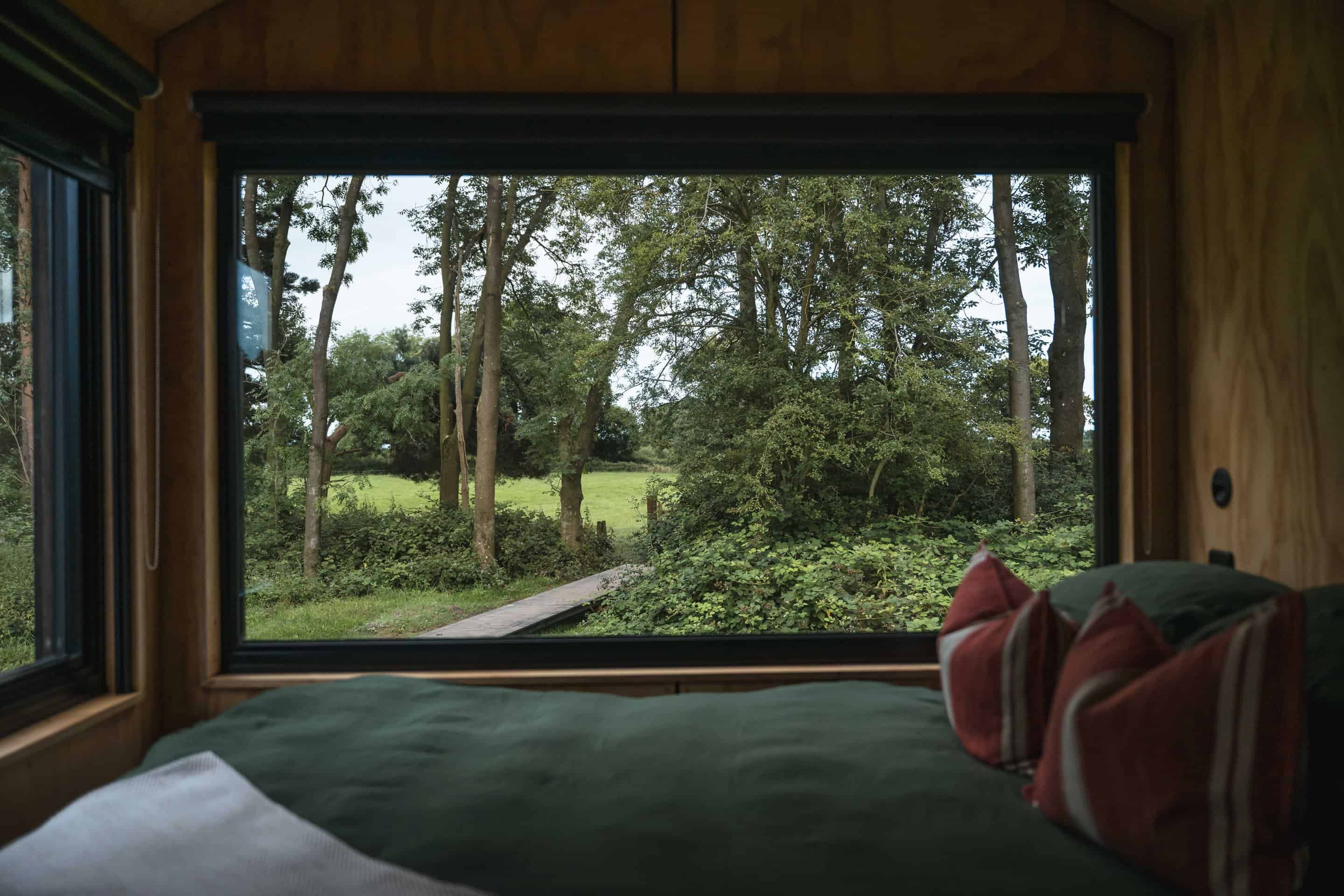 Wooden cabin interior overlooking lush green trees and pathway, eco-friendly off-grid living, nature retreat, secluded forest accommodation, sustainable off grid stay, outdoor adventure, connect with nature, eco tourism, green living, outdoor escape, wilderness retreat.