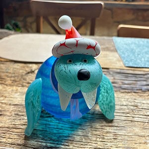 Glass figure of a walrus with a Christmas hat on a wooden table. The character named Wally wears white tusks, a scarf and radiates festive coziness.