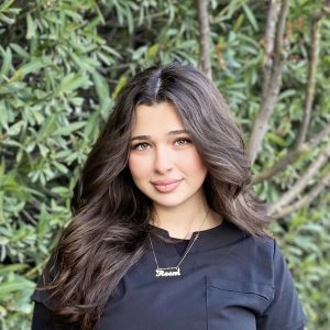A young woman with long, wavy dark hair and striking green eyes, wearing a personalized necklace, standing outdoors among lush greenery, representing brain performance and cognitive enhancement.