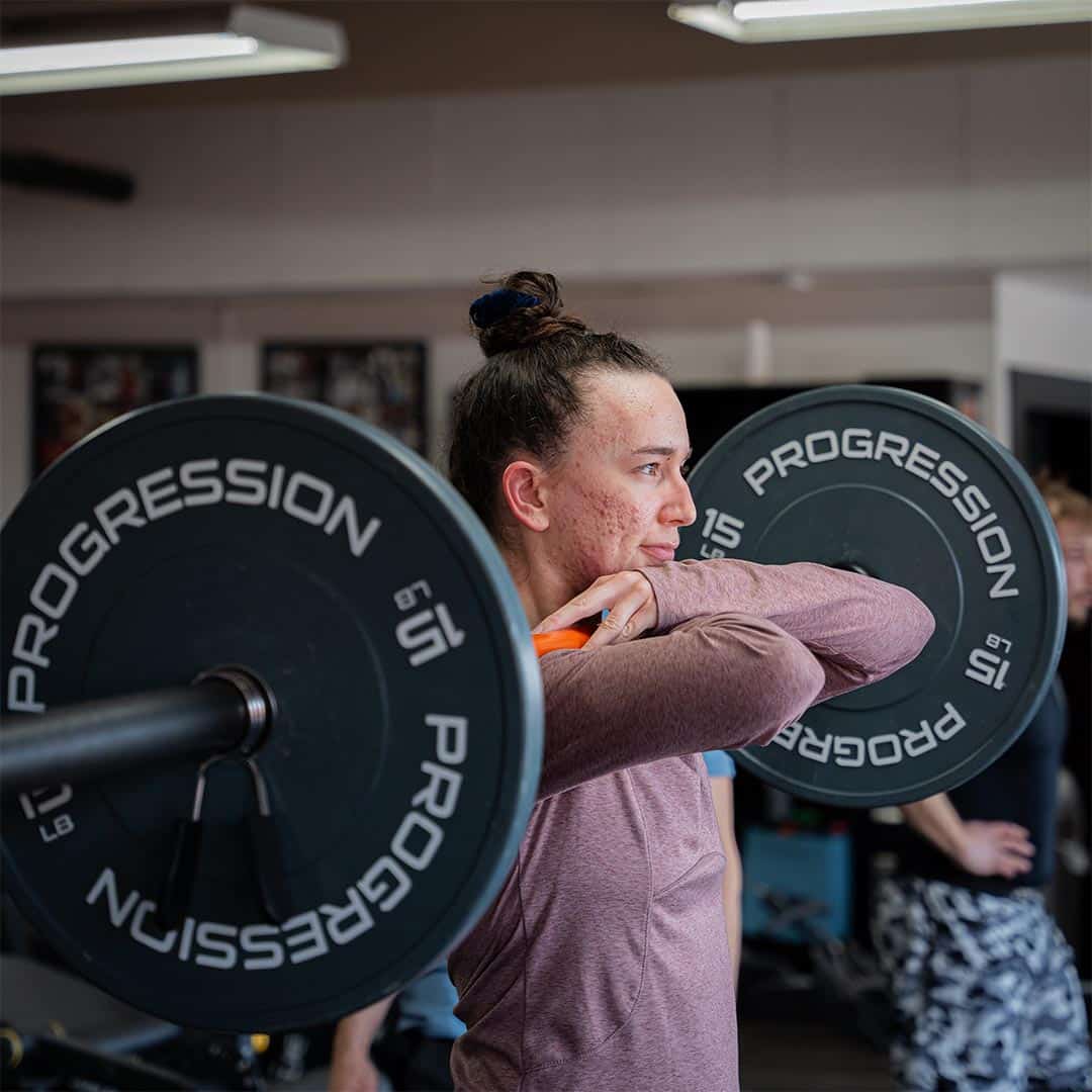 Heavy weightlifting woman executing shoulder press with barbell at Bravé MMA and Fitness gym.