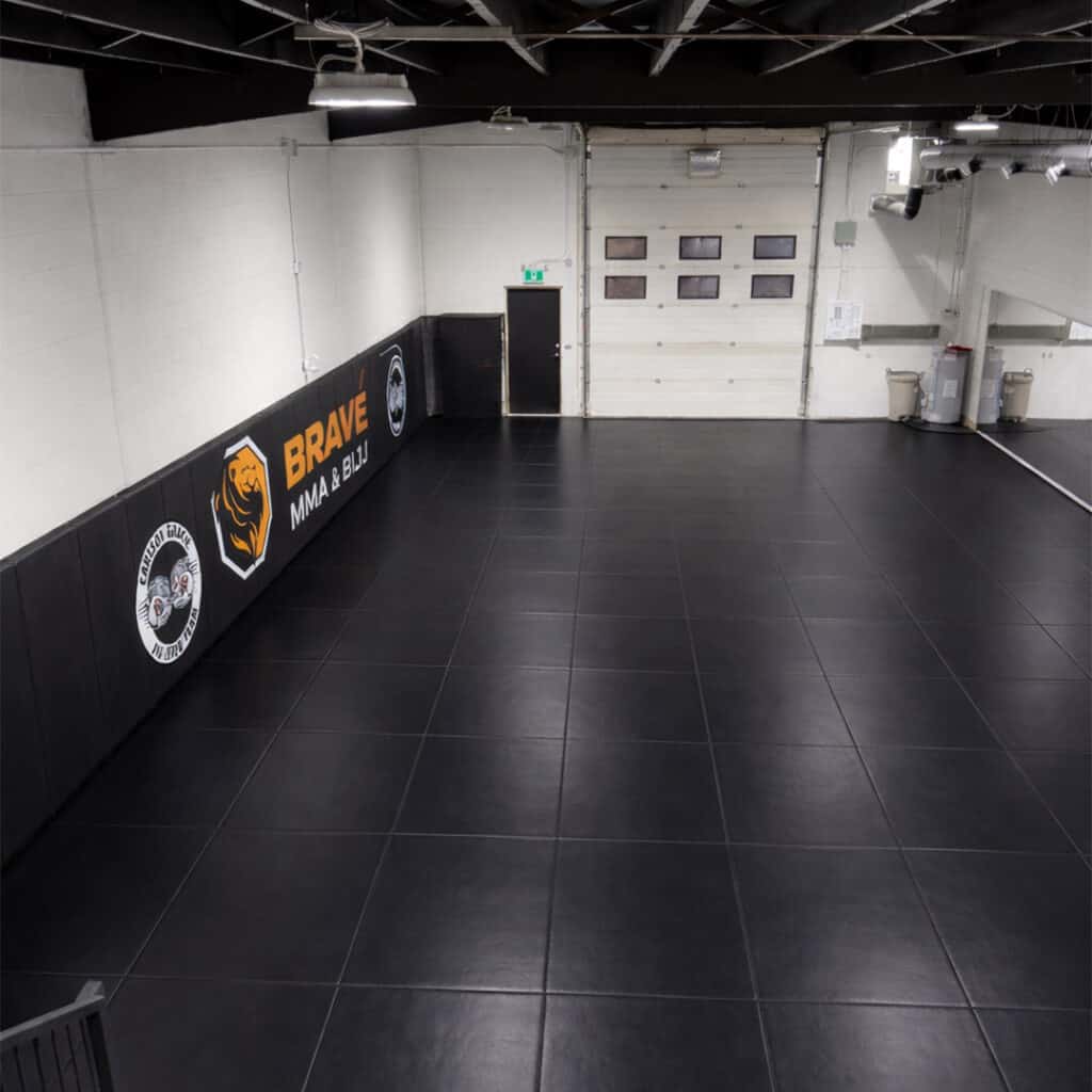 Bright, spacious MMA & BJJ training room with black mats, wall padding, and high ceilings. Features branding on walls, a large garage door, and industrial lighting, ideal for martial arts practice and competitions.