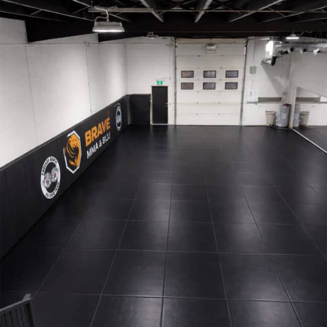 Bright, spacious MMA & BJJ training room with black mats, wall padding, and high ceilings. Features branding on walls, a large garage door, and industrial lighting, ideal for martial arts practice and competitions.