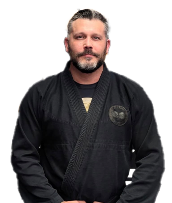 Male martial arts instructor with beard and slicked-back hair wearing a black gi, standing in front of a black and orange martial arts logo background.