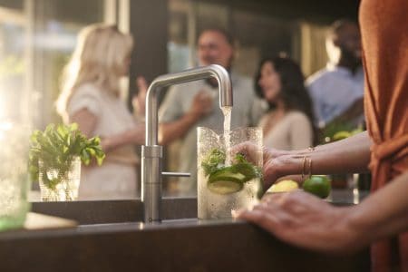 Sleek modern kitchen sink with fresh lime and herbs, contemporary tap, and a blurred group of people enjoying a social gathering in the background.