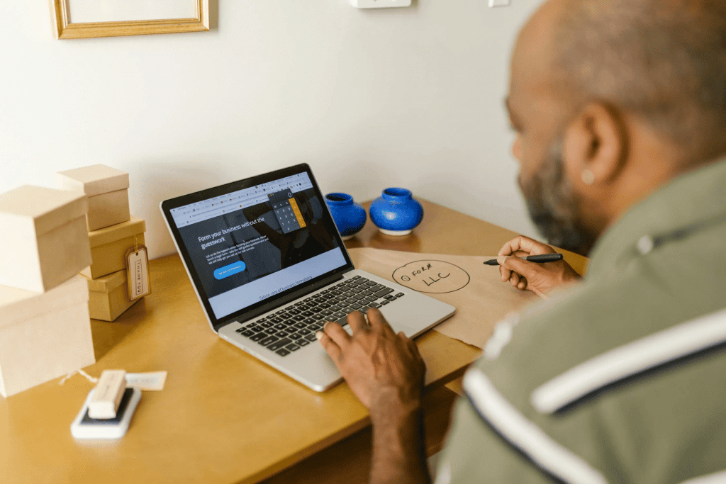 A man sitting at his computer, reviewing an LLC maintenance requirement while using a calculator app. The image represents the process of managing annual LLC fees, highlighting the importance of compliance and financial planning. With BusinessAnywhere, these tasks are automated, ensuring stress-free LLC maintenance.