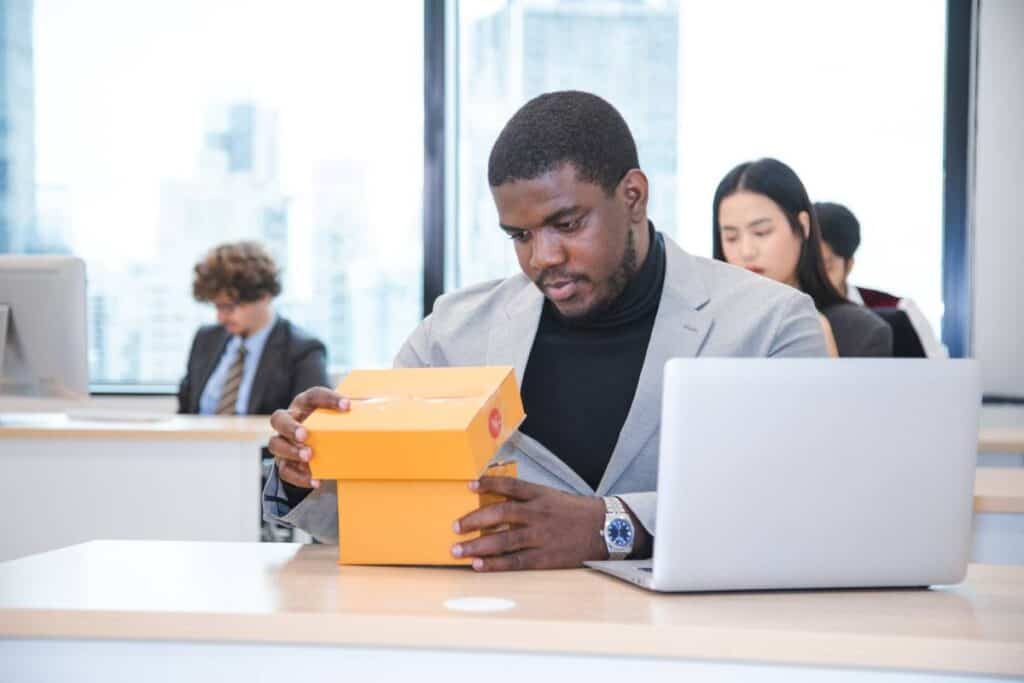 Man opening a package in an office setting with colleagues nearby.