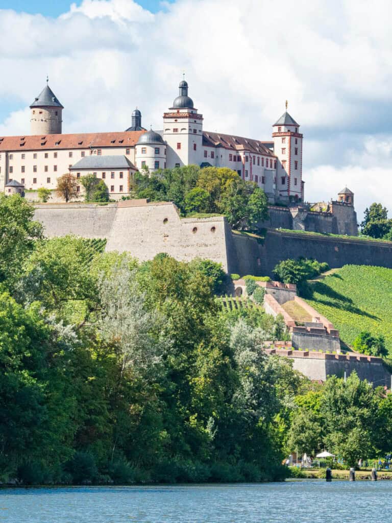 Die Festung Marienberg thront majestätisch über dem Main in Würzburg, umgeben von üppigem Grün und Weinbergen. Der helle Himmel mit weißen Wolken und das glitzernde Wasser im Vordergrund verstärken die malerische Schönheit der historischen Burganlage.