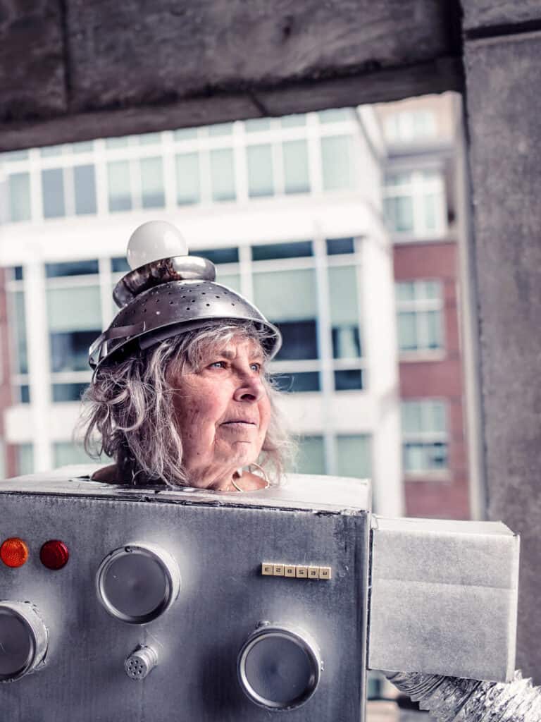 Ein älterer Mensch trägt ein kreatives Roboterkostüm aus Pappkarton, Dosen und einem Abtropfsieb mit Glühbirne auf dem Kopf, das futuristische und nostalgische Elemente kombiniert. Im Hintergrund ist ein modernes Stadtgebäude zu sehen, das einen spannenden Kontrast zur DIY-Ästhetik des Outfits bietet.