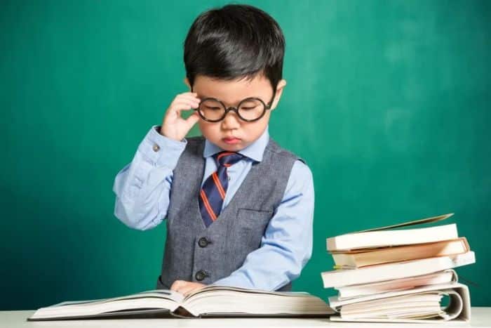 Bambino con occhiali e gilet legge un libro davanti a una lavagna verde