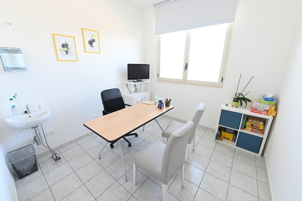 Ufficio luminoso con scrivania in legno, sedie grigie, lavandino a parete e scaffale con giochi e libri in uno spazio accogliente.