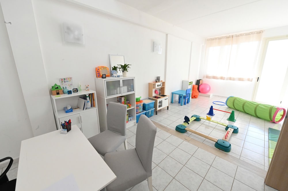Ampia sala luminosa con mobili bianchi, sedie grigie, scaffali pieni di giochi e attrezzature per attività motorie infantili.