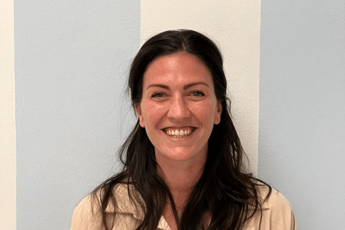 Donna sorridente con capelli lunghi scuri davanti a una parete con strisce verticali bianche e azzurre.